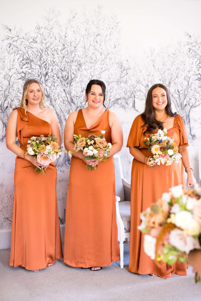 Bridesmaid in satin rust coloured dresses holding bouquets of flowers in autumn shades