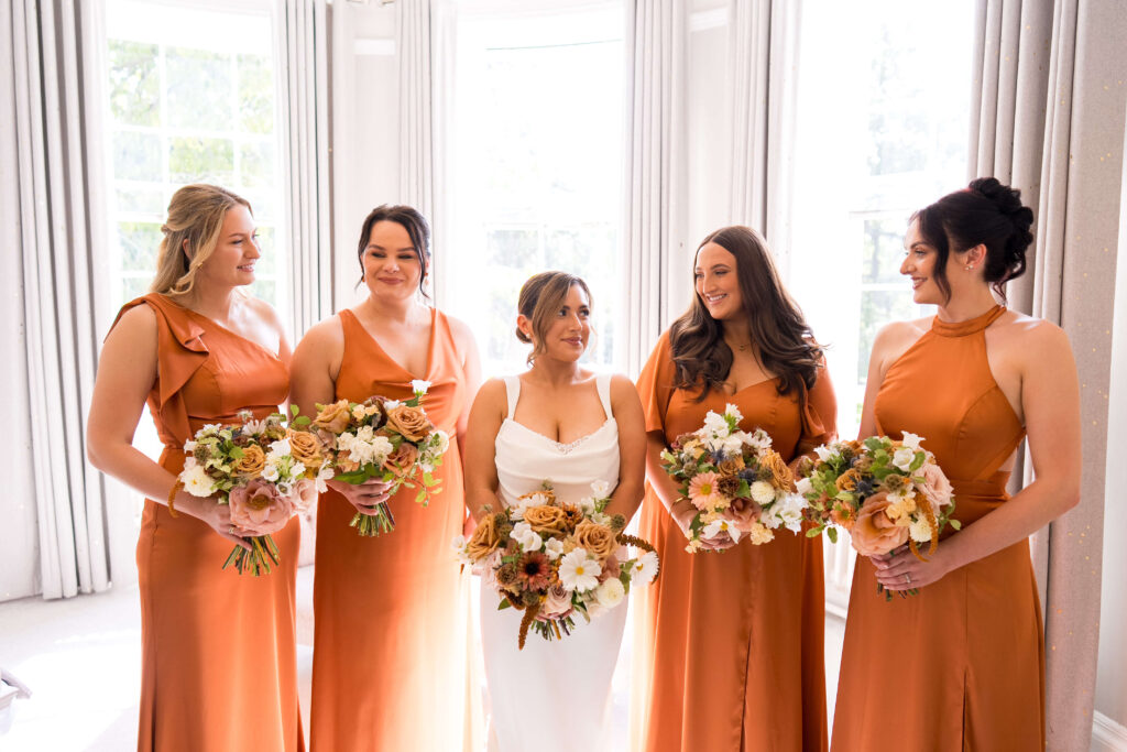 Bridesmaid in satin rust coloured dresses holding bouquets of flowers in autumn shades
